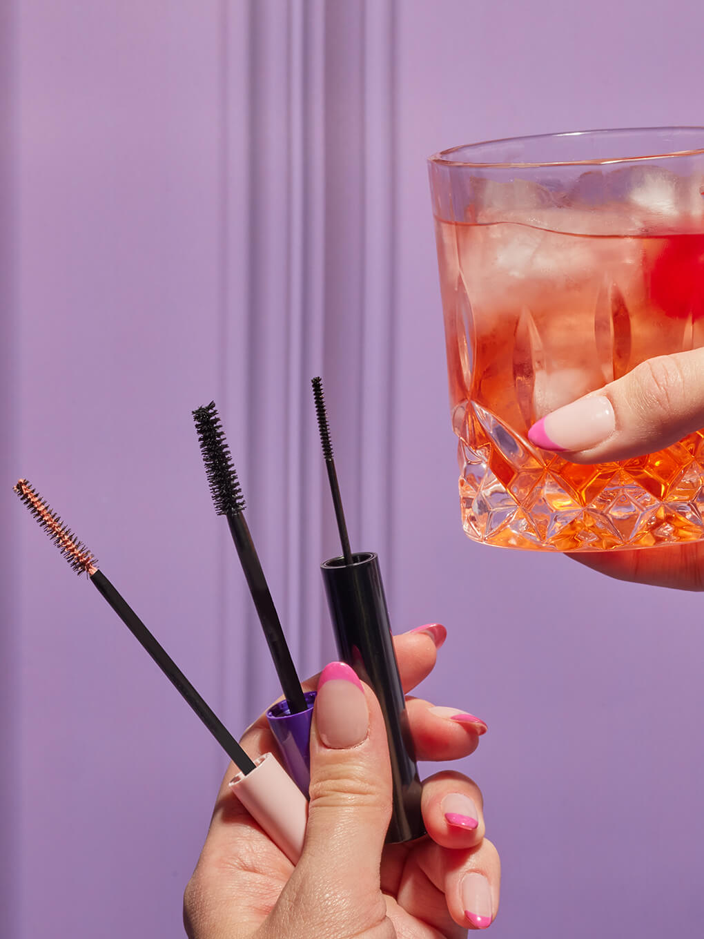 hand holding up two mascaras and a lash primer next to cocktail