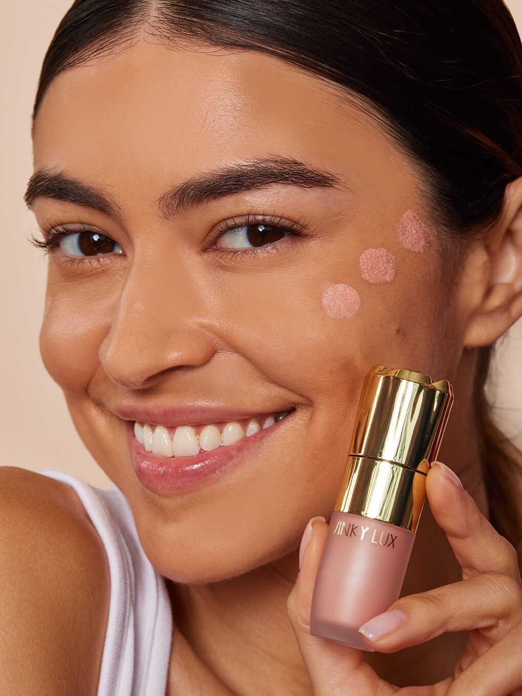 brunette model smiling holding up cheeky rose liquid highlighter with product dotted on cheek