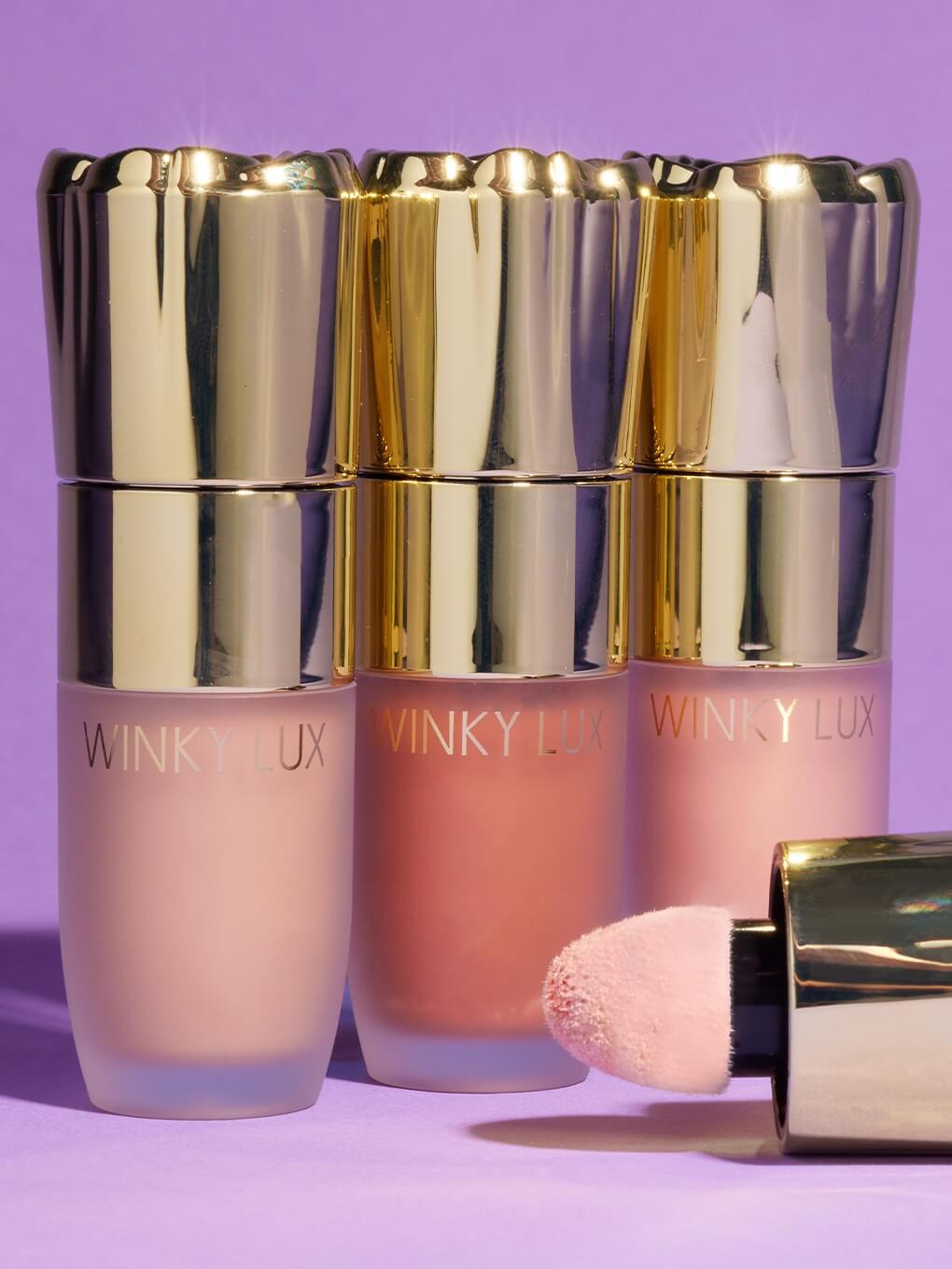 three shades of cheeky rose liquid highlighter next to each other on purple background with cap lying in front of