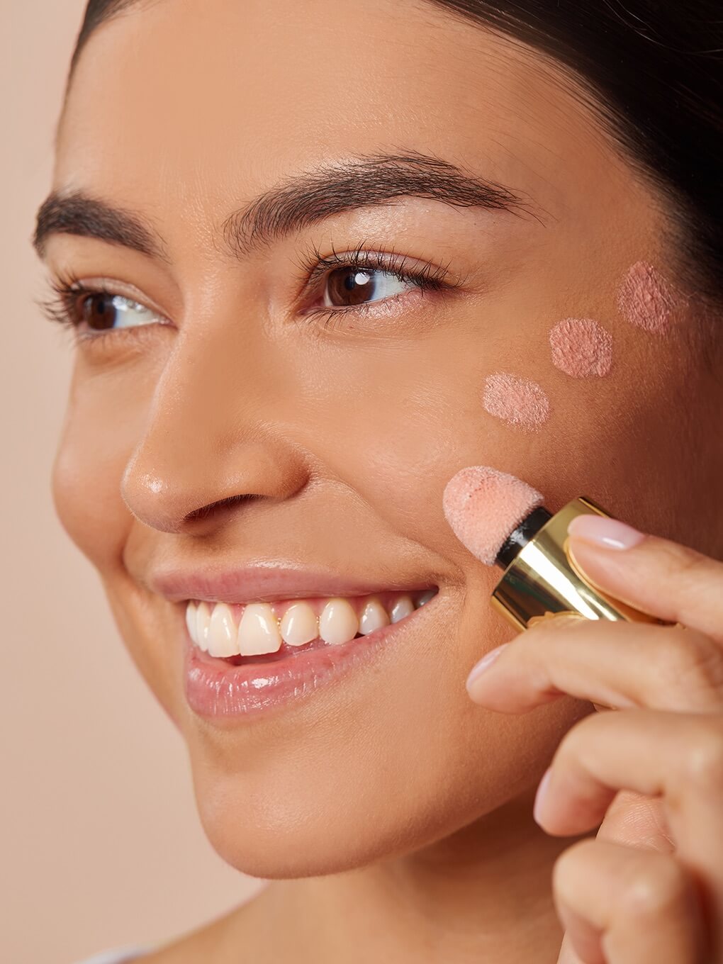 Angel -- model smiling applying dots of cheeky rose liquid highlighter to high cheekbone