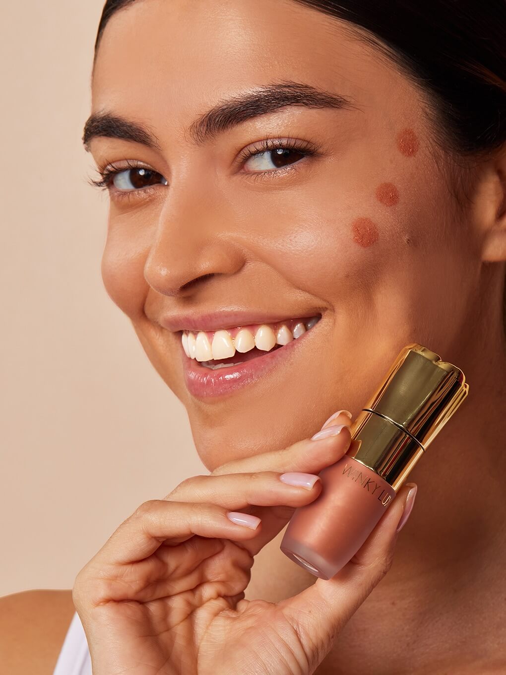 Nova -- model smiling holding up cheeky rose liquid highlighter next to cheek